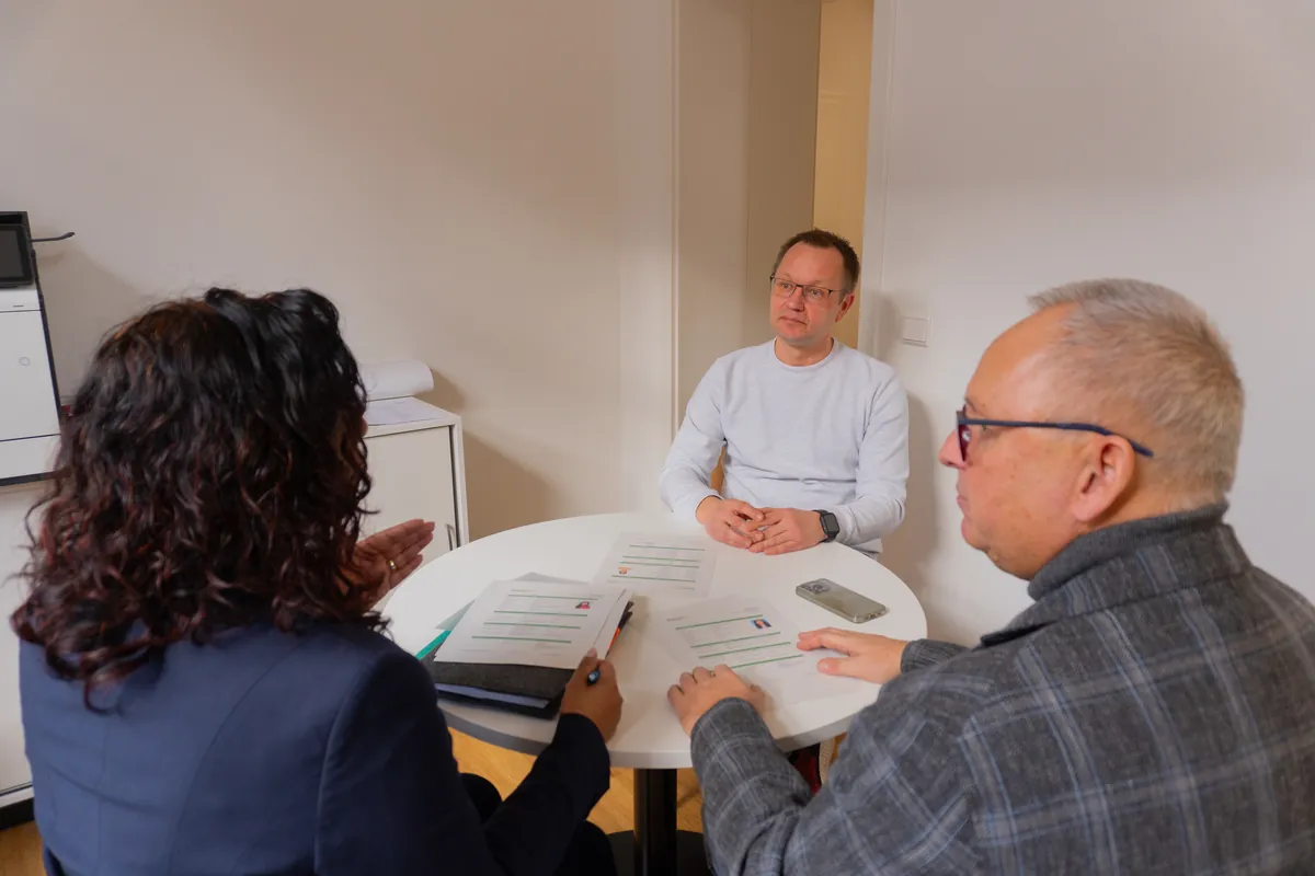 Baicy Terbrüggen im Gespräch mit zwei Männern am runden Besprechungstisch, auf dem Unterlagen liegen
